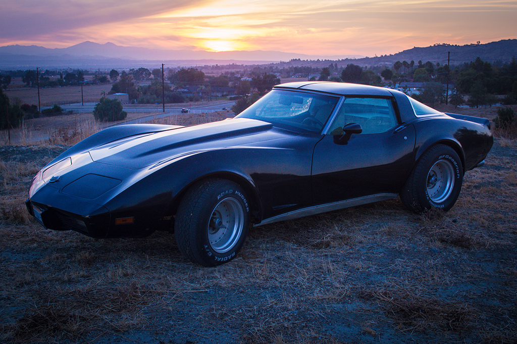 A classic American muscle car rests at sunset, its sleek lines and deep silhouette set against a glowing horizon and open countryside.